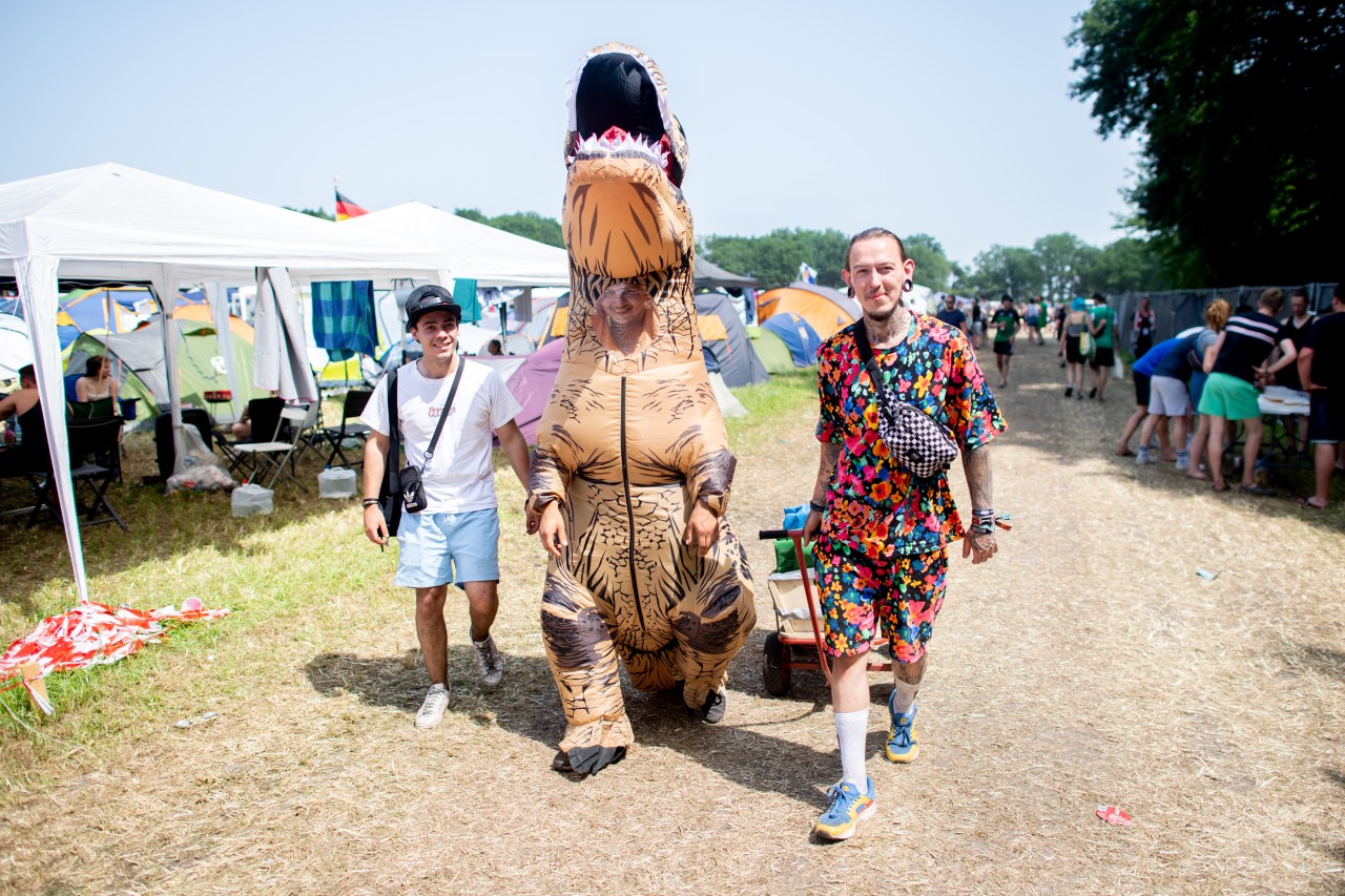Dennis geht, als T-Rex verkleidet, mit seinen Freunden Christian (l) und Paul (r) über den Zeltplatz vom Hurrican Festival.